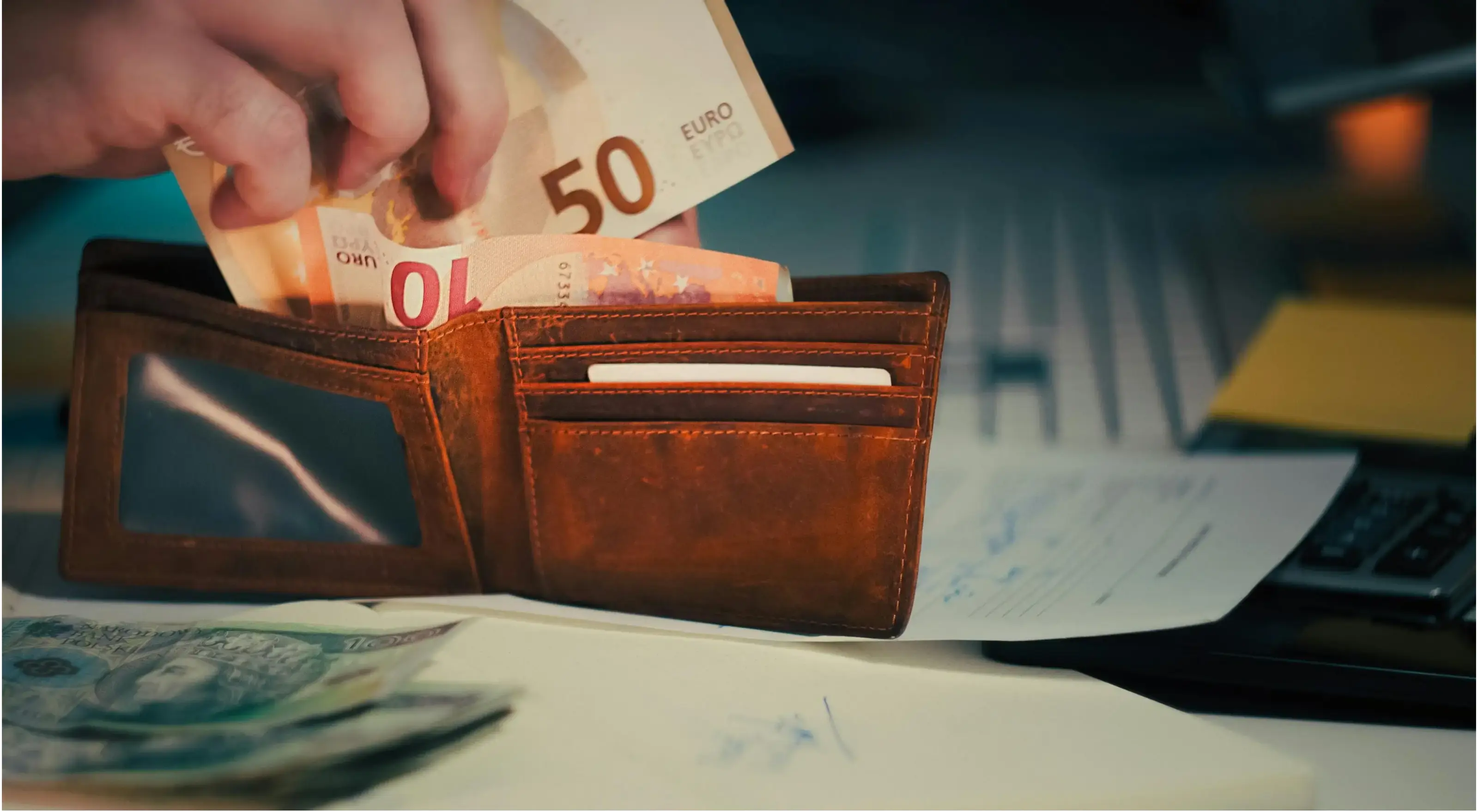 Hand die eurobiljetten in een bruine leren portemonnee stopt, met documenten en een rekenmachine op de achtergrond.