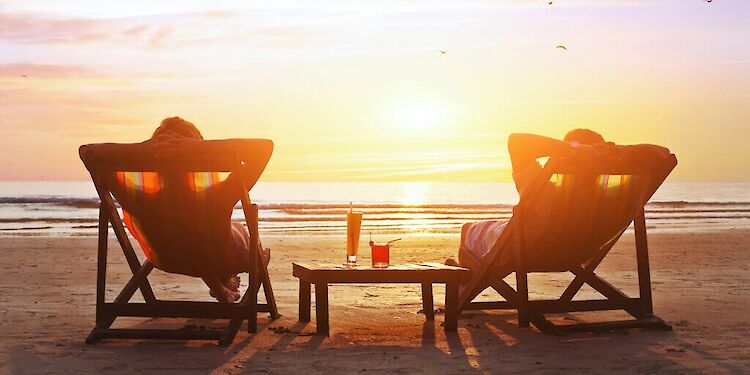 twee mensen op strandbedjes met een tafel met cocktails tussen hun in kijkend naar de zon die ondergaat