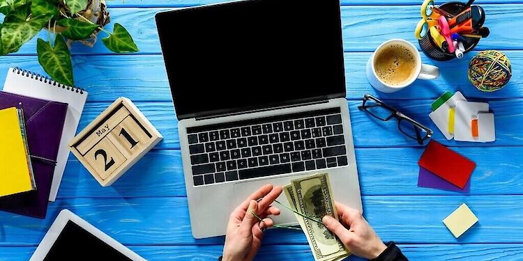 bureaublad met laptop, koffie, datum, notitieboekjes en 2 handen met geld erin.