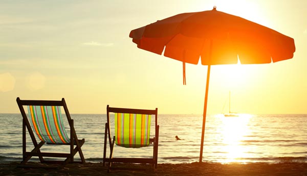 twee strandstoeltjes met een parasol ernaast met de zee en een zon die ondergaat op de achtergrond