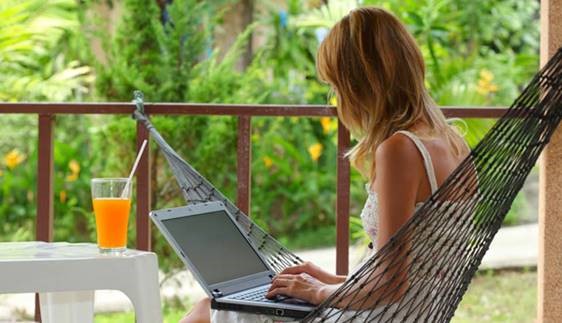 Vrouw zit in hangmat en tikt op laptop. Op de voorgrond staat een tafel met een cocktail daarop.