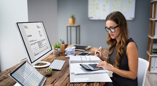 vrouw achter bureau met laptop, tablet en een map met een rekenmachine waar ze iets op uitrekent