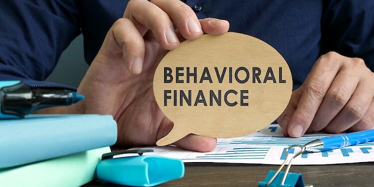 Een hand houdt een houten denkwolk vast waar 'Behavioral Finance' op staat geschreven