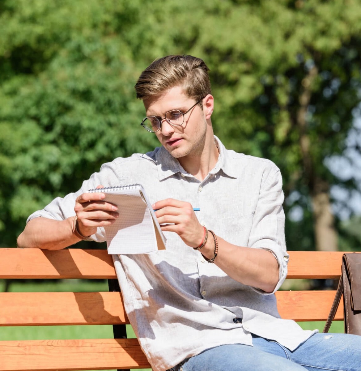 man met bril met notitieboekje en pen in de hand zittend op een bankje in het park